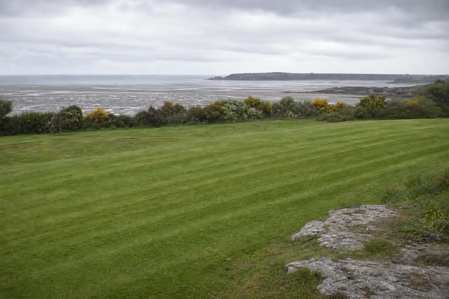 Grande pelouse fraîchement tondue en périphérie de Saint-Brieuc, bandes de coupe nettes et régulières, horizon ouvert sur la baie sous une lumière côtière ventée, haie littorale en arrière-plan et sol de schiste affleurant.