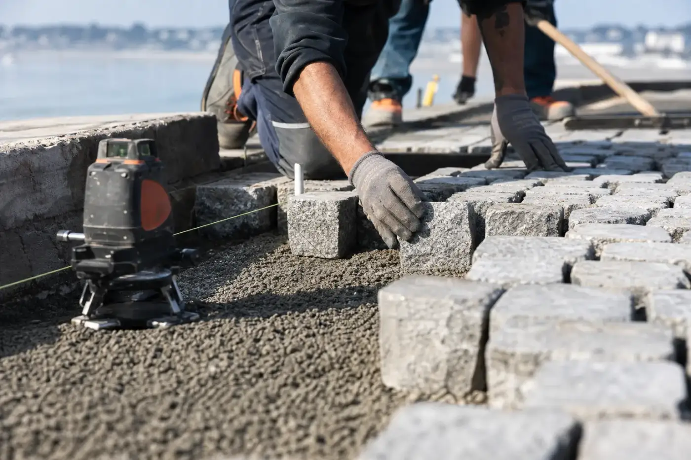 Gros plan sur la pose de pavés de granit sur lit de graves compactées, niveau laser réglant la pente, chantier de bord de mer.