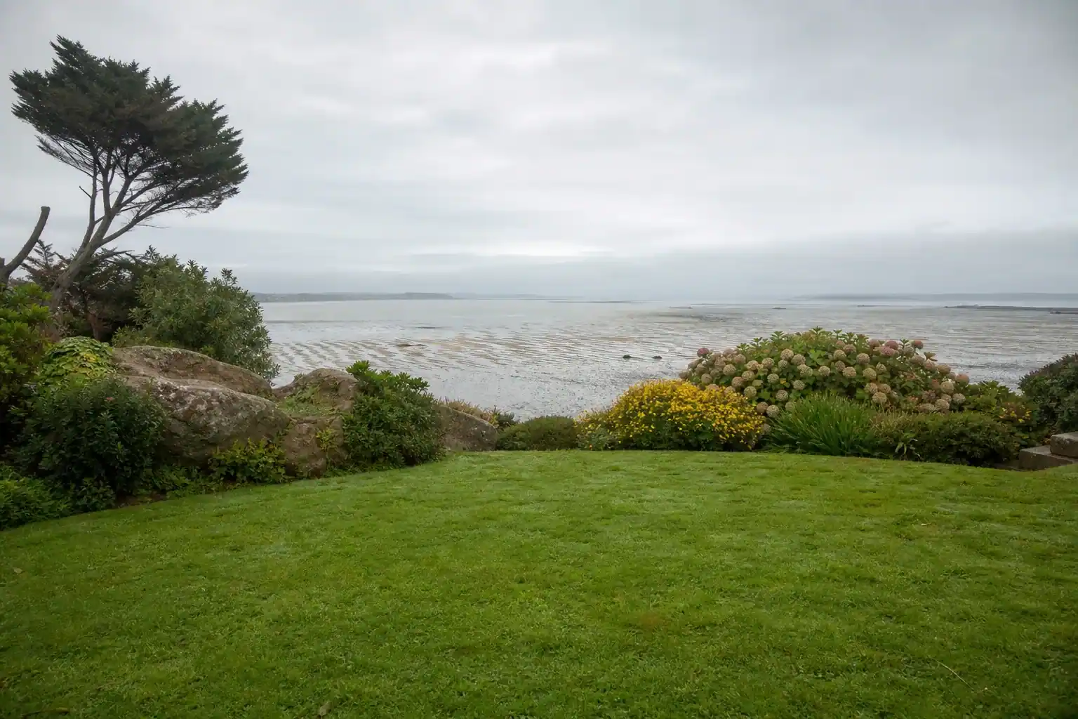 Jardin de particulier à Langueux ouvert vers le fond de baie, pelouse fraîche et massif bas résistant au vent, lumière humide de bord d'estran, grèves découvertes devinées au loin, cadrage large et horizontal au ras du sol.