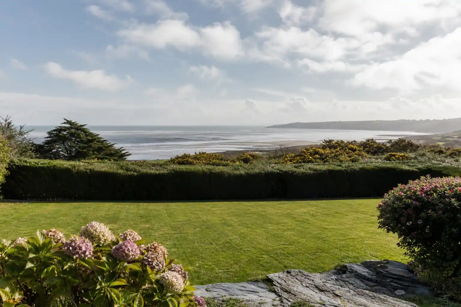 Jardin pavillonnaire sur les hauteurs de Saint-Brieuc, pelouse fraîchement tondue et haie taillée nette, horizon ouvert sur la baie et la Manche, lumière côtière lumineuse et ventée, sol de schiste affleurant en bordure.